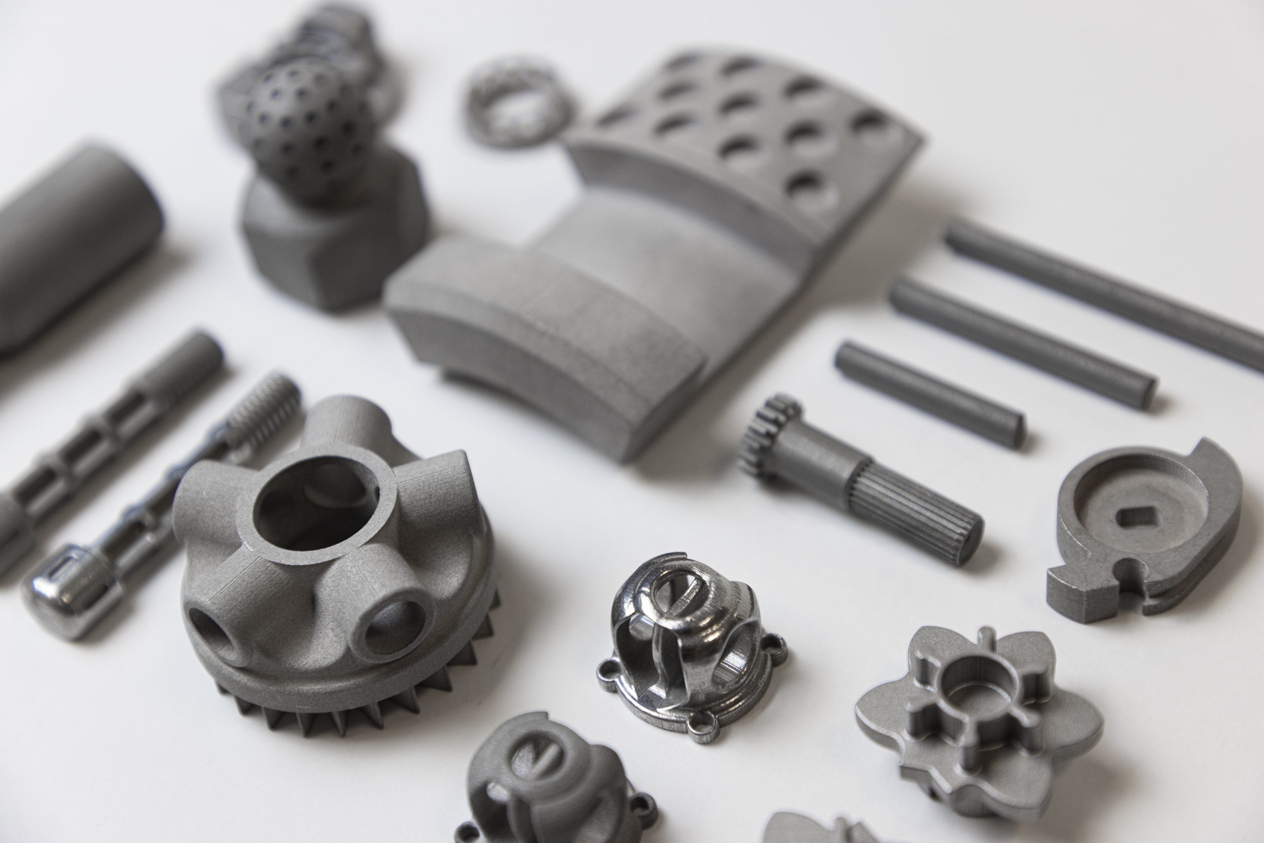 An array of high-precision, matte grey 3D printed metal and polymer industrial parts, showcasing complex geometries and fine details on a clean white background.